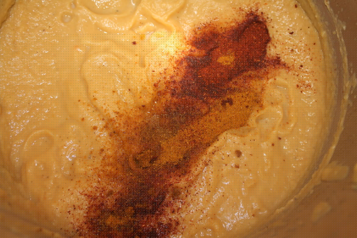 A closely zoomed image of a stainless steel pan, filled with curried parsnip soup, with an array of colourful mixed spices on the surface.